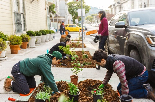 Community members kneeling on the sidewalk planting plants in a row of beds that have been installed in the concrete.