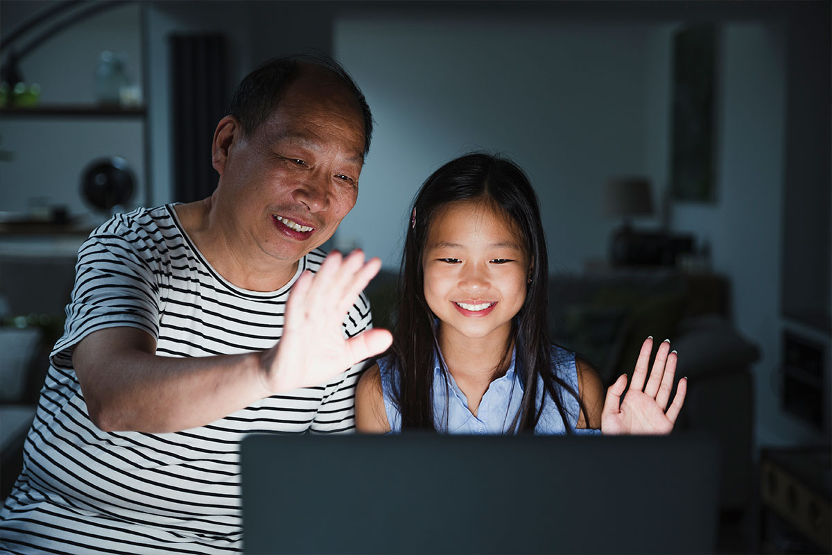 Un abuelo y una nieta sonríen y saludan a alguien en la pantalla de una computadora por la noche